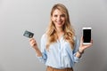 Happy young pretty blonde business woman posing grey wall background holding credit card showing display of mobile phone