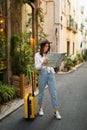 Happy young pretty european woman tourist in hat looking for hotel on map with suitcase in city