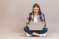 Happy young woman sitting on the floor with crossed legs and using laptop isolated on gray background Royalty Free Stock Photo