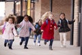 Happy young school girls wearing coats and carrying schoolbags running in a walkway with their classmates outside their infant sch
