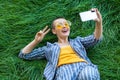 Happy young short hair woman in casual blue striped suit, yellow shirt, glasses lying down on green grass with mobile phone, doing Royalty Free Stock Photo