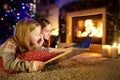 Happy young sisters reading a story book together by a fireplace in a cozy dark living room on Christmas eve. Celebrating Xmas at