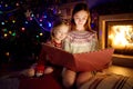 Happy young sisters reading a story book together by a fireplace in a cozy dark living room on Christmas eve. Celebrating Xmas at