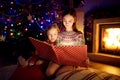 Happy young sisters reading a story book together by a fireplace in a cozy dark living room on Christmas eve. Celebrating Xmas at