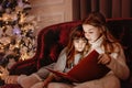 Happy young sisters reading a story book together by a fireplace in a cozy dark living room on Christmas eve