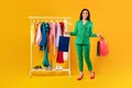 Happy young stylish european woman posing with shopper bags near clothing rail, satisfied with great sales