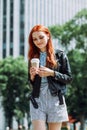 Happy young trendy red hair woman drinking take away coffee and walking in an urban city. Royalty Free Stock Photo