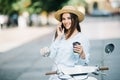 Happy young woman drinking takeaway coffee near her moped and talking phone on city street Royalty Free Stock Photo