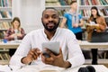 Happy young university multiracial students studying with books in library. Handsome African guy looking at camera while Royalty Free Stock Photo