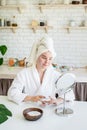 Happy young woman applying face scrub on her face in her home kitchen looking at the mirror Royalty Free Stock Photo