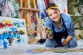 Happy young woman in apron smiling at camera, holding paintbrush while working on painting, sitting on the floor at home Royalty Free Stock Photo