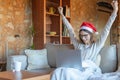 Happy young woman with arms outstretched glasses and Santa hat sitting on the sofa at home