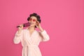 Happy young woman in bathrobe with hair curlers and sunglasses singing into hairbrush on pink background, space for text Royalty Free Stock Photo