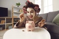 Happy young woman in beauty face mask and hair curlers putting coins in piggy bank Royalty Free Stock Photo