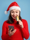 Happy young woman in a Christmas cap and red sweater holds a smartphone and a credit card. On blue