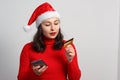 Happy young woman in a Christmas cap and red sweater holds a smartphone and a credit card. On white