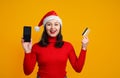 Happy young woman in a Christmas cap and red sweater holds a smartphone and a credit card. On a yellow background