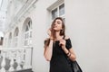 Happy young woman with cute smile in a stylish black dress with a leather fashionable backpack enjoys a walk around the city on a Royalty Free Stock Photo