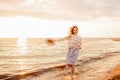 Happy young woman in a dress holding straw hat and walking alone on empty sand beach at sunset sea shore and smiling. Freedoom, Royalty Free Stock Photo