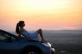 Happy young woman driver in blue dress enjoying warm summer evening laying on her car hood. Travelling and vacation concept Royalty Free Stock Photo