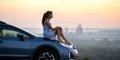 Happy young woman driver in blue dress enjoying warm summer evening sitting on her car hood. Travelling and vacation concept Royalty Free Stock Photo