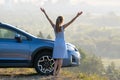 Happy young woman driver with outstretched up hands enjoying warm summer evening standing beside her car. Travelling and vacation Royalty Free Stock Photo