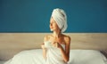 Happy young woman enjoying with cup of coffee drying her wet hair after bath with wrapped towel on her head sitting on bed looking Royalty Free Stock Photo