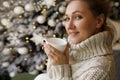 Happy young woman enjoying drinking tea or coffee in living room with Christmas decor over new year tree background Royalty Free Stock Photo
