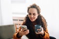 Happy young woman enjoying mobile technology while drinking coffee at home. Smiling lady using smartphone for online shopping and Royalty Free Stock Photo