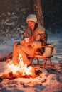 Happy young woman in warm knitted hat and jacket sits on wooden sled by bonfire with cup of hot tea