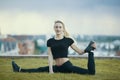 Happy young woman on the grass performs twine with the bent leg, on background cityscape Royalty Free Stock Photo