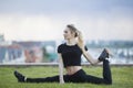 Happy young woman on the grass performs twine with the bent leg, on background cityscape Royalty Free Stock Photo