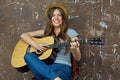 Happy young woman with hat sitting with acoustic guitar on grunge wall.