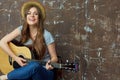 Happy young woman with hat sitting with acoustic guitar on grunge wall. Royalty Free Stock Photo