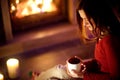 Happy young woman having a cup of hot chocolate by a fireplace in a cozy dark living room on Christmas eve. Celebrating Xmas at