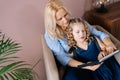 Happy young woman and her little curly daughter reading book together while sitting on armchair Royalty Free Stock Photo