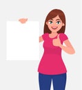 Happy young woman holding a blank / empty sheet of white paper or board and gesturing thumbs up sign.