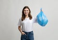 Happy young woman holding blue plastic garbage bag, standing on grey background