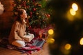 Side view of happy woman holding gift box while having video call on laptop near Christmas tree. Royalty Free Stock Photo