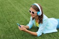 Happy young woman holding phone listening to music in headphones lying on grass in summer park Royalty Free Stock Photo