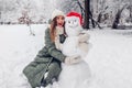 Happy young woman hugs snowman in snowy winter park dressed in Christmas Santa hat. Festive fun outdoor activities Royalty Free Stock Photo