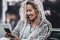 Happy young woman in knitted woolen jacket and curly hair text messaging using mobile phone while sitting outdoors on bench. Happy Royalty Free Stock Photo