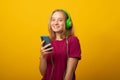 Happy young woman listens music in headphones holds mobile phone chooses song from playlist dressed in casual t-shirt Royalty Free Stock Photo