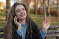 Happy young woman with long wavy brown hair sits on bench in park talks on phone and gesticulating. Girl in autumn park Royalty Free Stock Photo