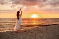 Happy young woman in a long white dress looking at the sunset on empty sand beach with her hands up. Freedoom, vacation, Royalty Free Stock Photo