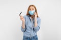 Happy young woman in a medical mask on her face, showing a plastic credit card and talking on a mobile phone, on a white