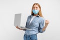 Happy young woman in a medical mask on her face, uses a laptop and shows a winning gesture, on a white background