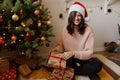 Happy young woman opening christmas gift box and smiling under christmas tree with lights in modern room. Stylish girl in santa Royalty Free Stock Photo