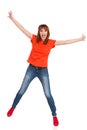 Happy young woman in orange shirt, jeans and red sneakers is standing on one leg, holding arms outstretched and shouting. Full Royalty Free Stock Photo