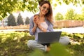 Happy young woman with paper cup of coffee and laptop sitting on green grass in park Royalty Free Stock Photo